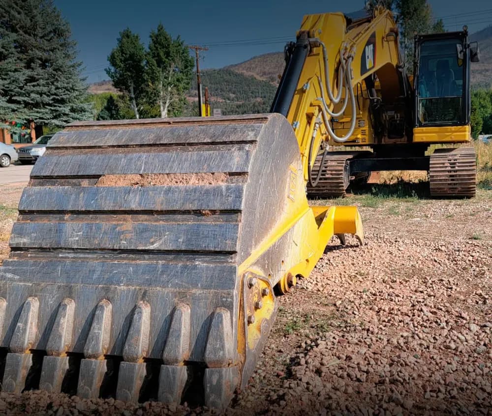A yellow excavator with its bucket resting on gravel at a mountain job site.