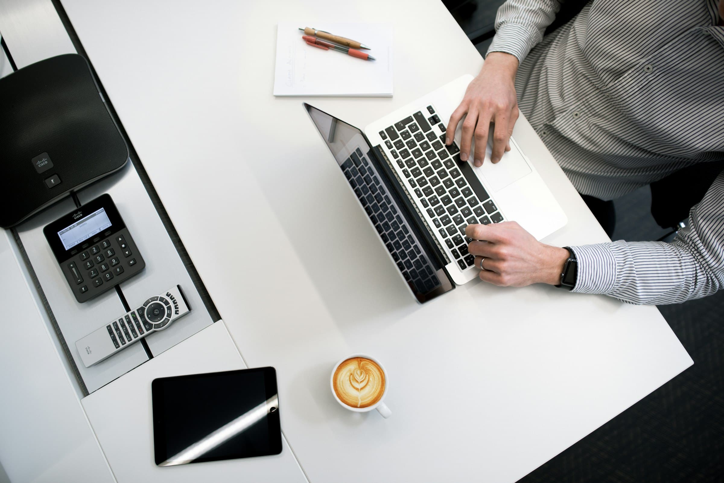 Overhead view of a person working on a laptop at a white desk with coffee and a notepad